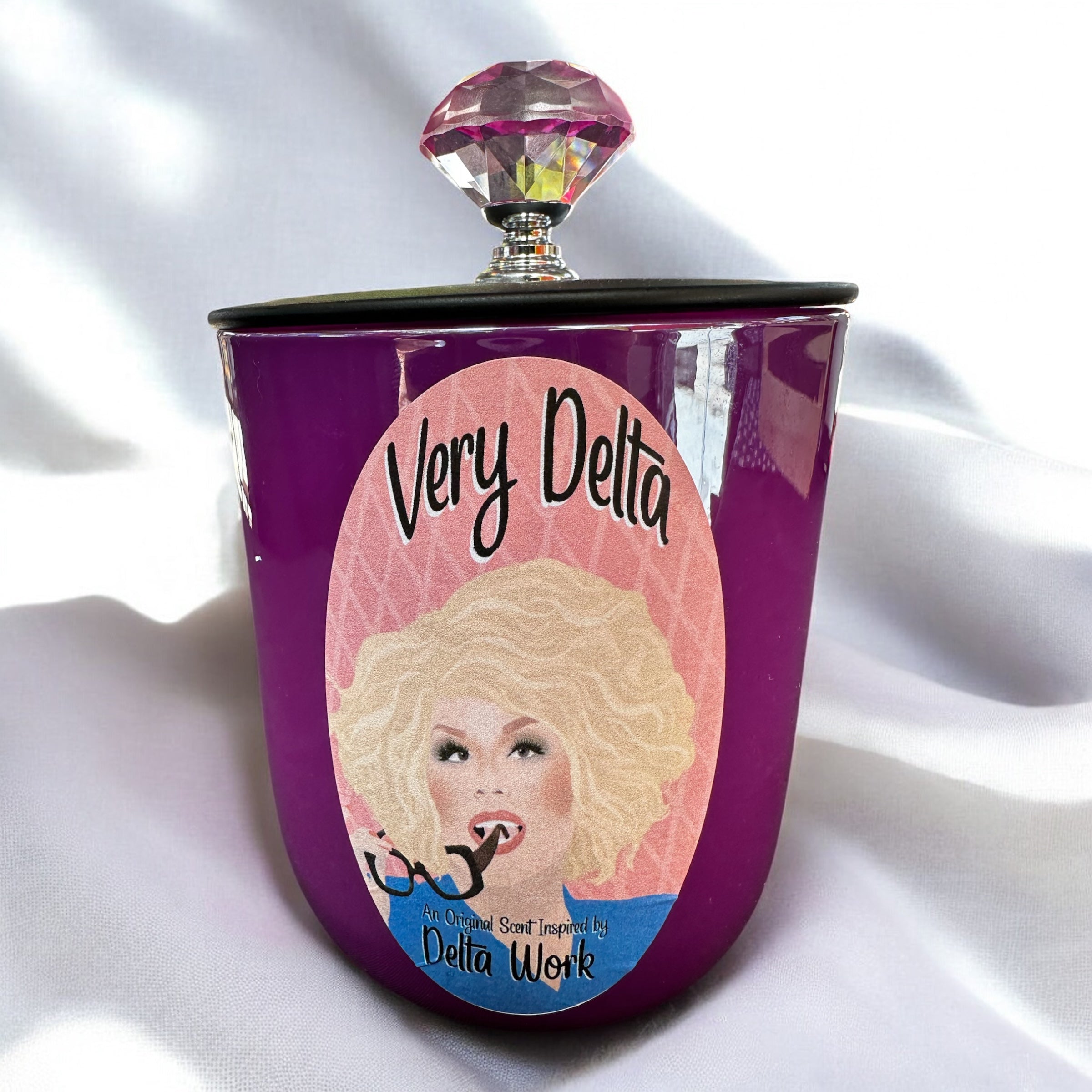 Purple candle with a pink lid featuring Delta Work and 'Very Delta' text on a white fabric background