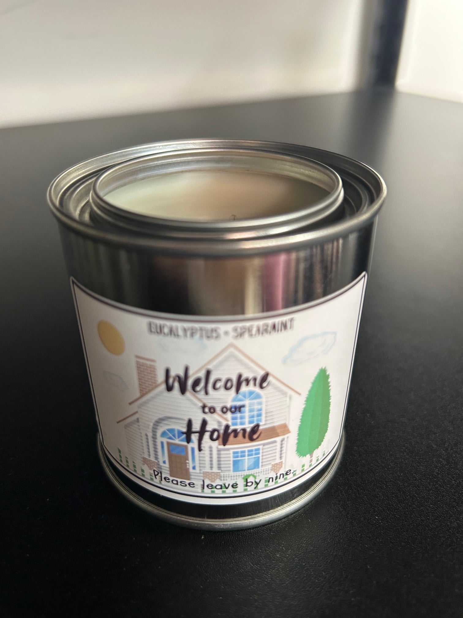 A candle with a label that reads 'Welcome to Our Home, Please Leave by 9', with an illustration of a house and trees.