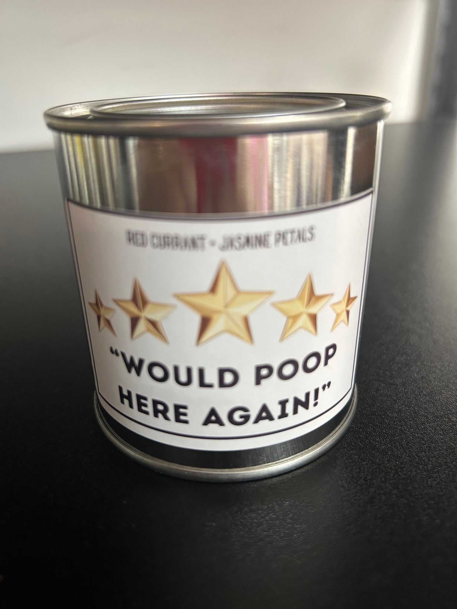 A candle with a label that reads 'Would Poop Here Again - 5 Stars' with a background featuring red currant and jasmine petals.