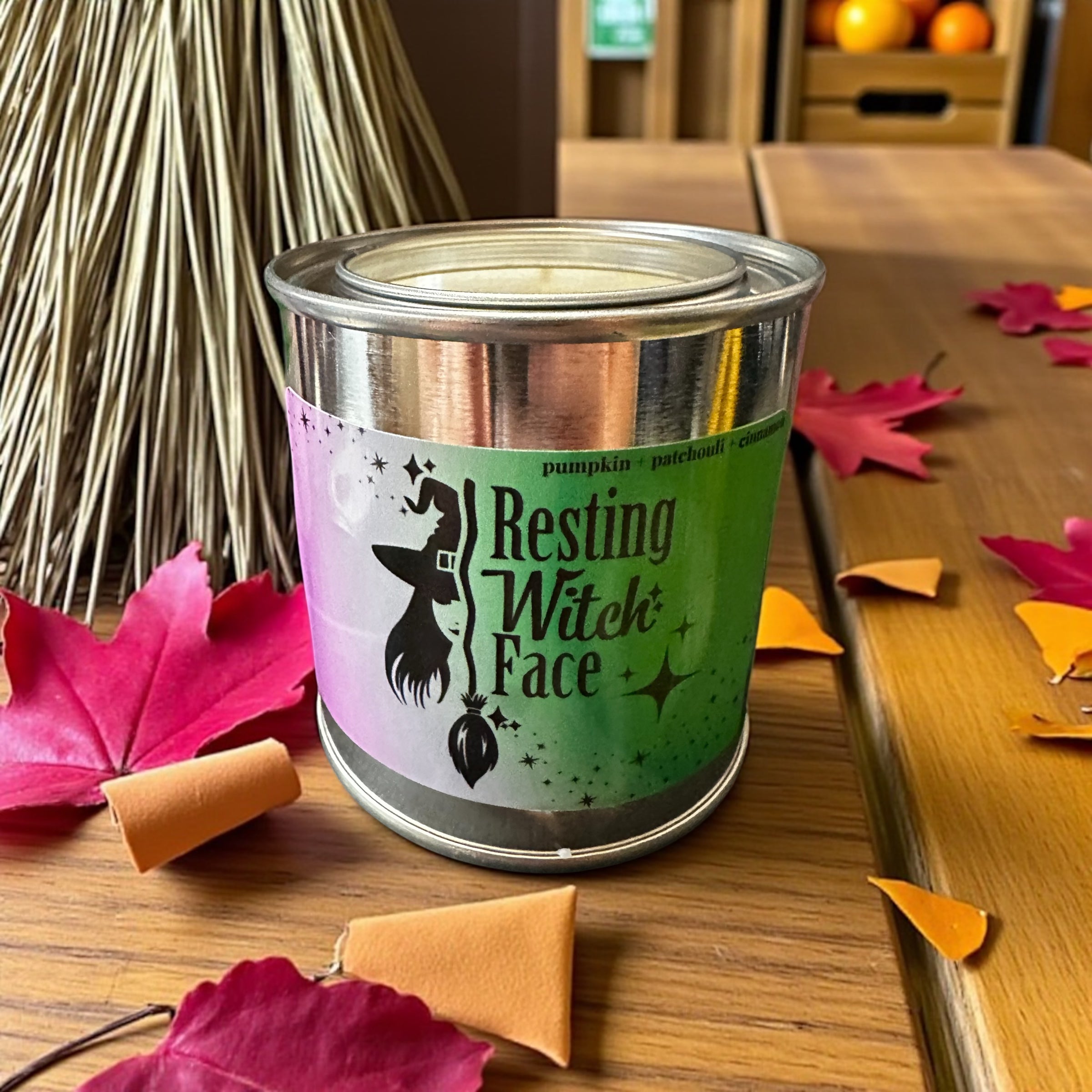 Candle in a can labeled 'Resting Witch Face' on a wooden surface with fall leaves.