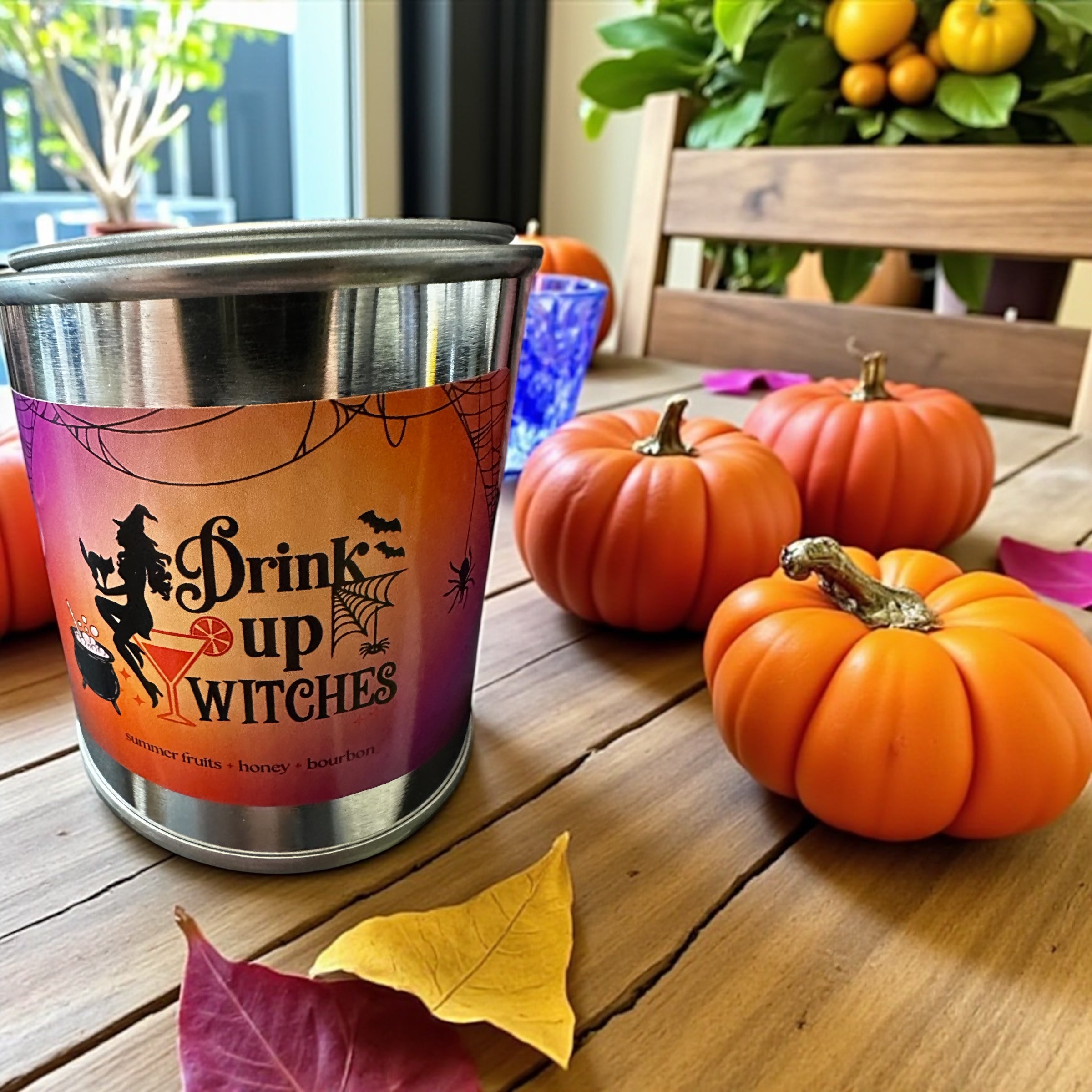 Stainless steel paint can candle with Halloween-themed label on a table with pumpkins and leaves.