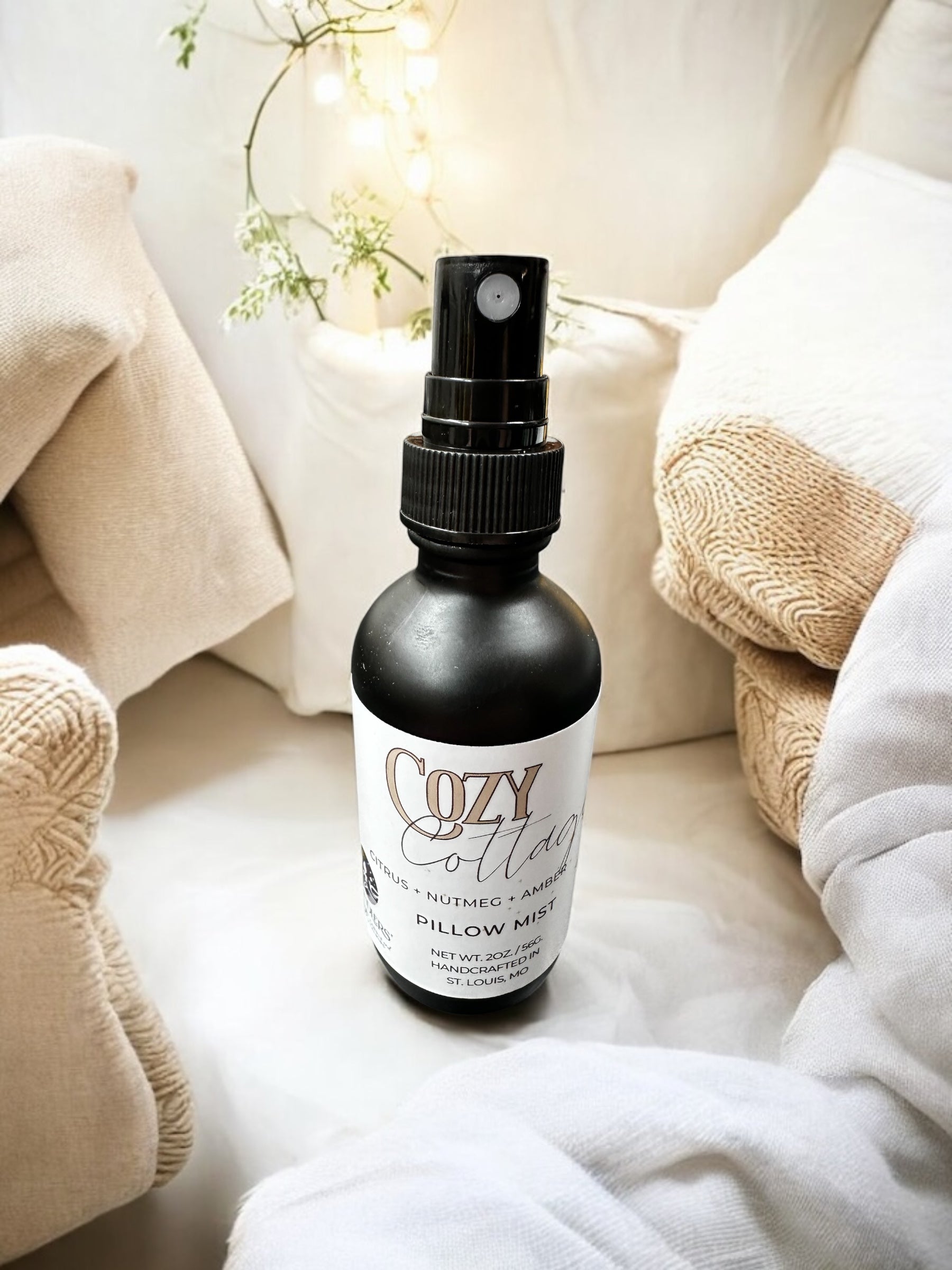A black spray bottle labeled 'Cozy Cottage Pillow Spray' placed on a bed with beige bedding, next to a white and green plant.
