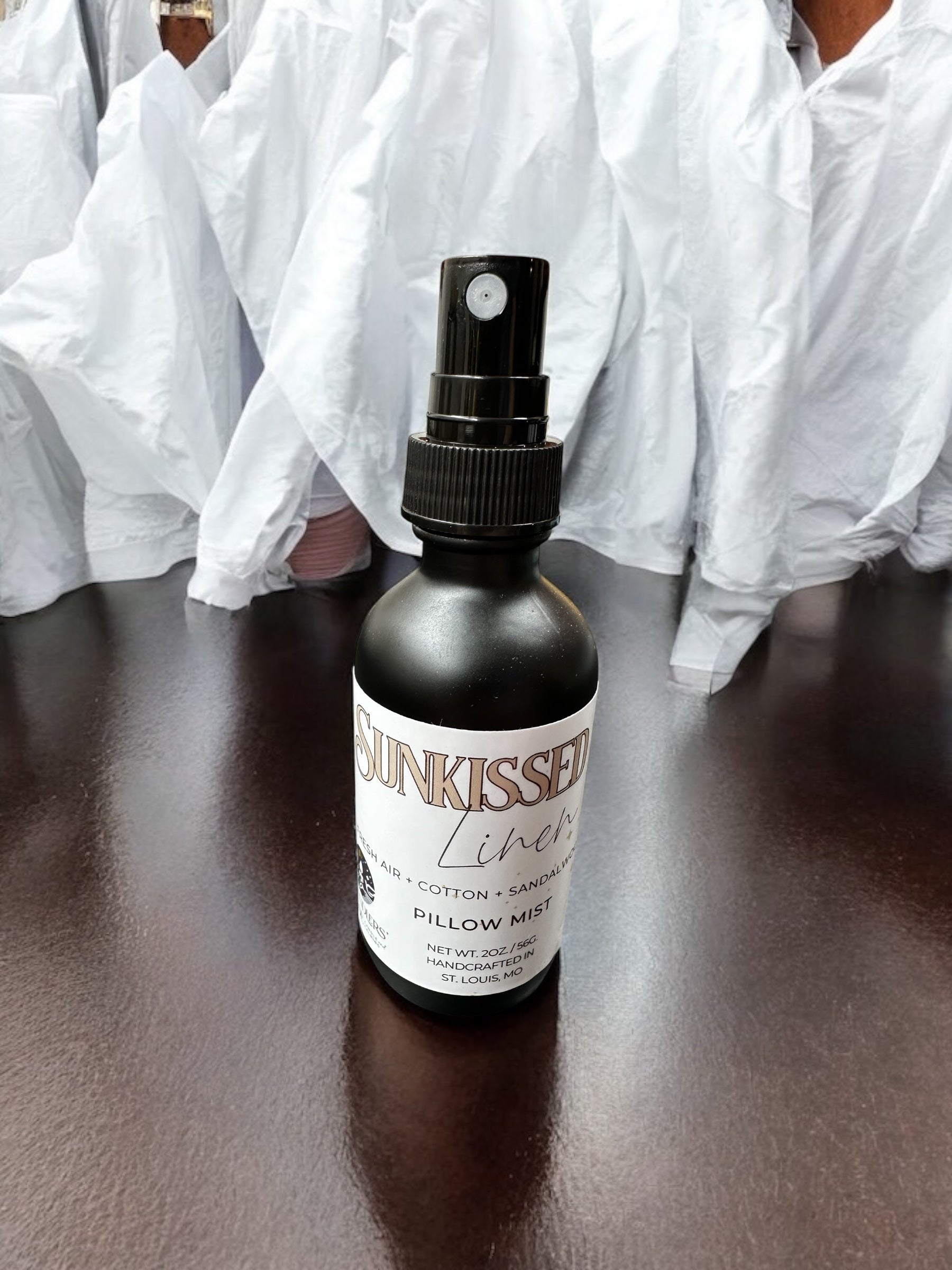 A black spray bottle labeled 'SUNKISSED LINEN' placed on a wooden surface with white linen in the background.