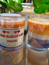 Two candles with clear glass bases, one closed and the other with a label that reads 'This Smells Like Pride!', placed on a reflective surface with a green plant in the background.
