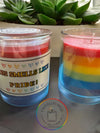 Two candles with multicolored layers in blue, red, and purple, placed on a table with a plant in the background. The candles have labels supporting LGBTQ+ rights, and the brand Chandler's Pride is visible.