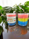 Two candles with multicolored layers in shades of red, blue, and green, placed on a surface with a green plant in the background and a logo in the foreground.