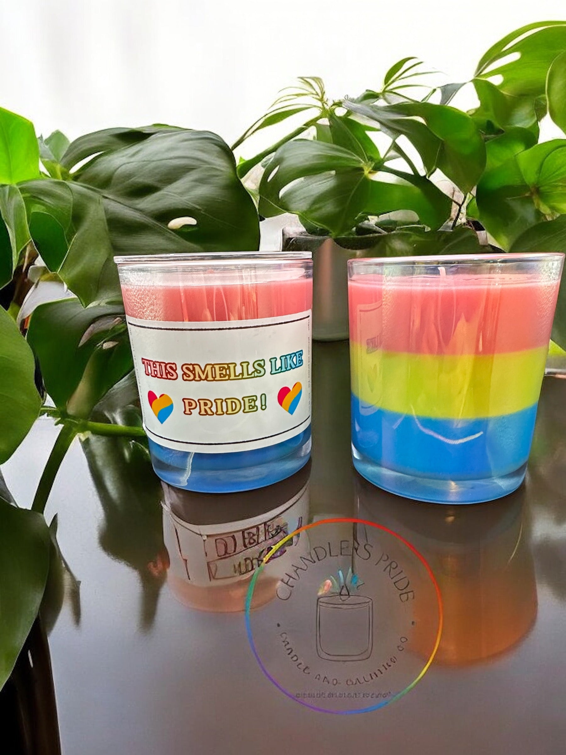 Two candles with multicolored layers in shades of red, blue, and green, placed on a surface with a green plant in the background and a logo in the foreground.