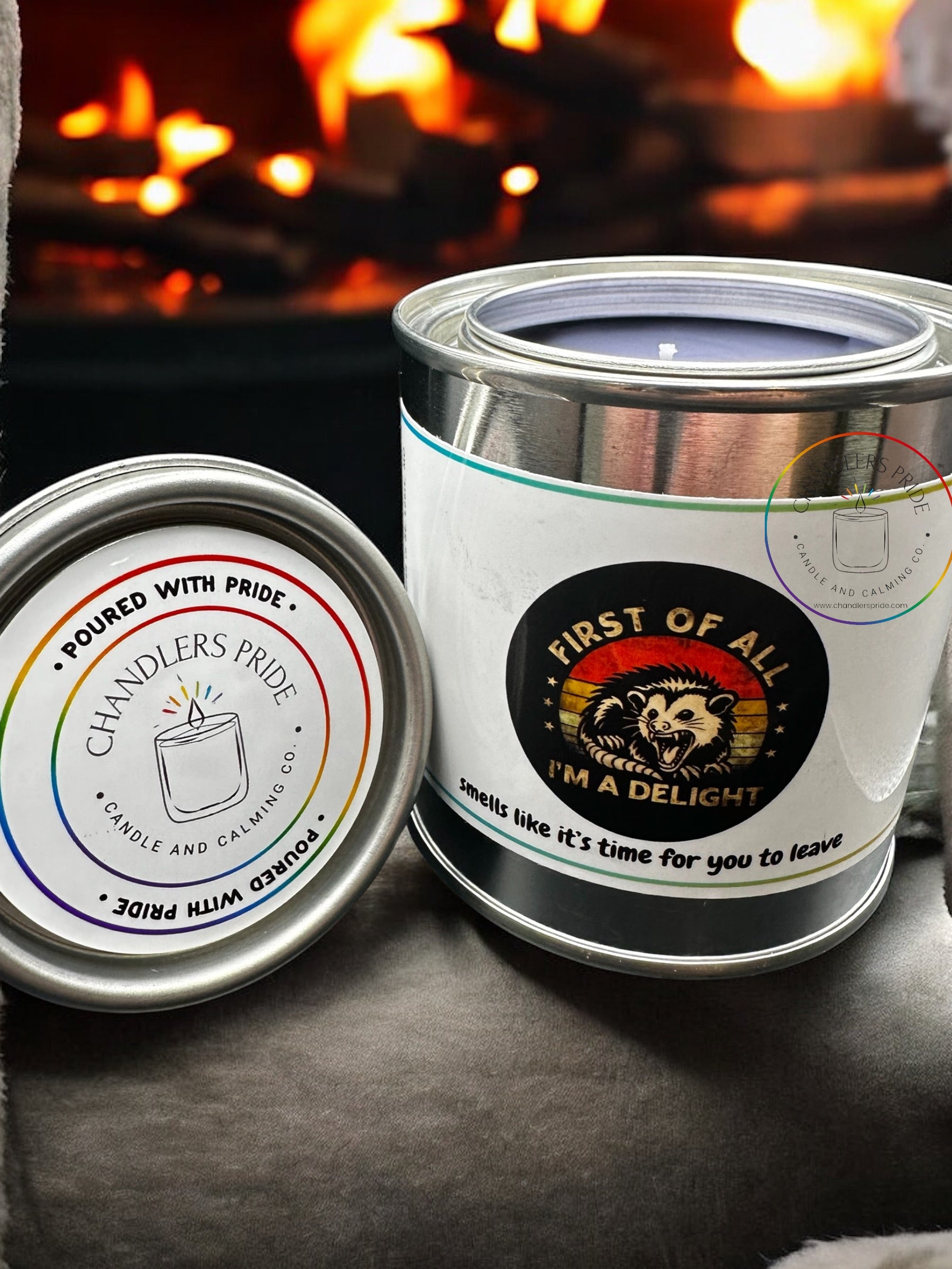 A set of two metal candle cans with colorful labels. One can is labeled 'Chandler's Pride' with a rainbow and stars design, and the other can is labeled 'First of All, I'm a Delight - Smells Like Time for You to Leave' with a more subdued design.