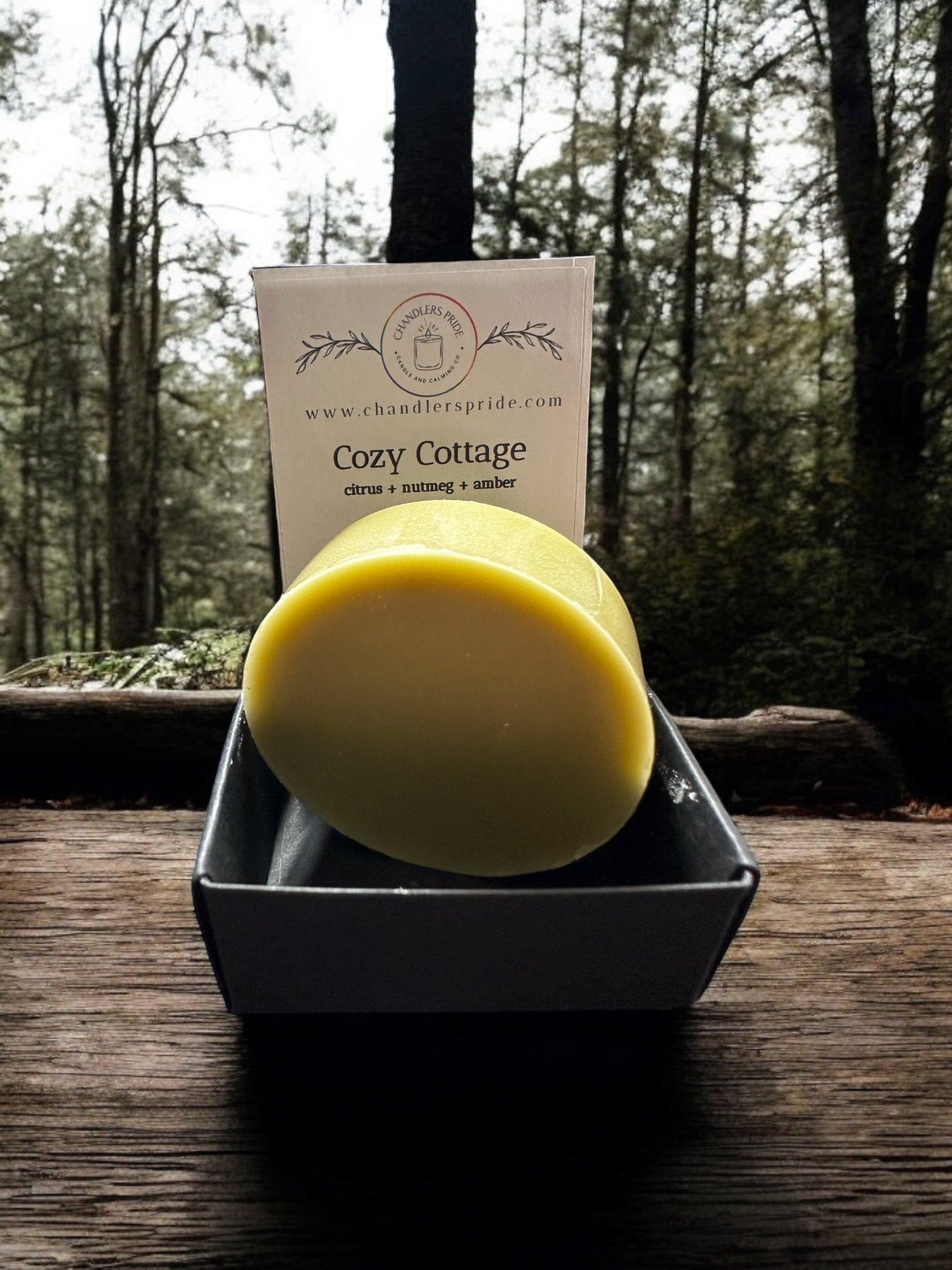 A black spray bottle labeled 'Cozy Cottage Pillow Spray' placed on a bed with beige bedding, next to a white and green plant.