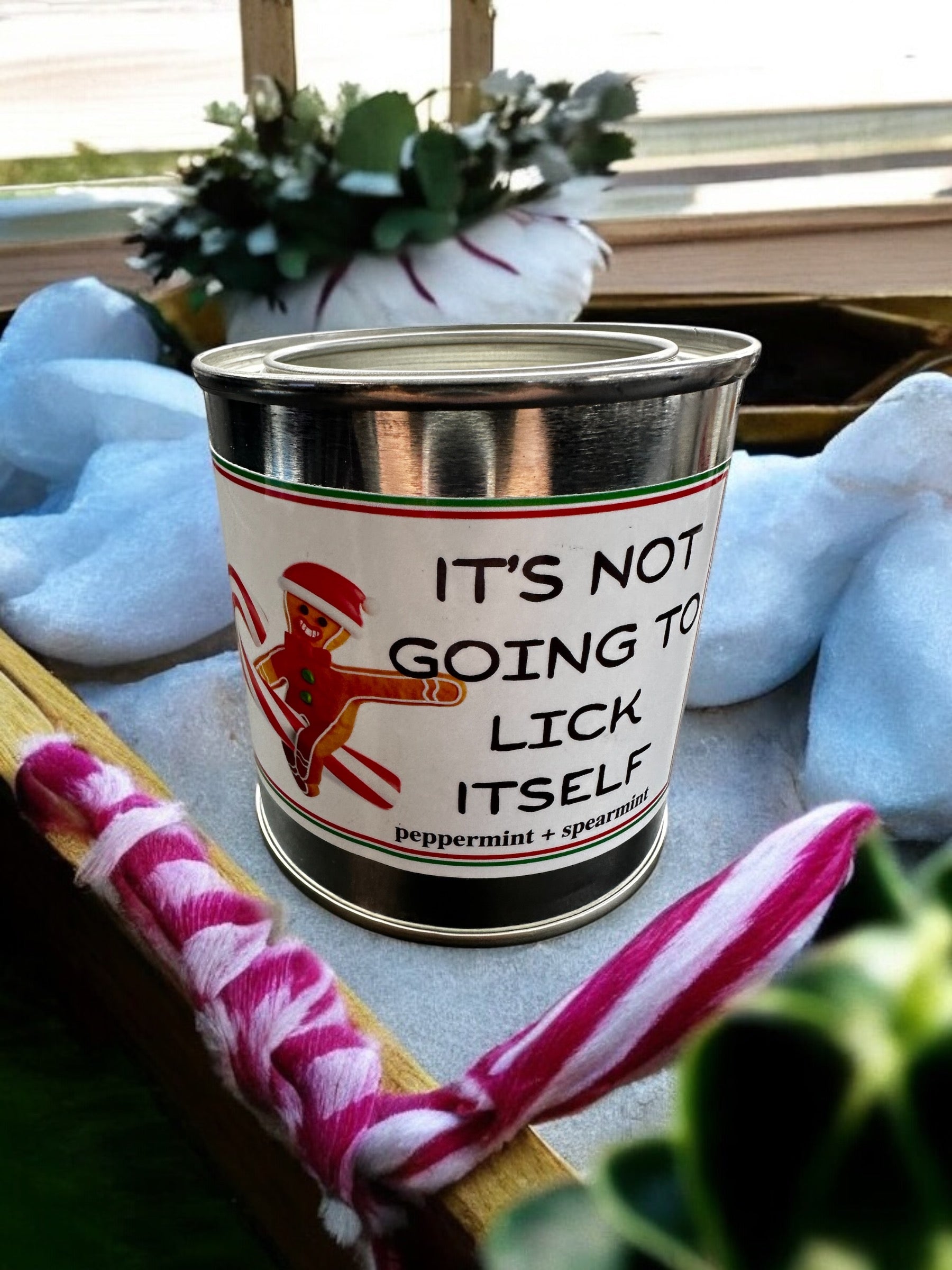 A candle with a label reading 'It's Not Going to Lick Itself', with a peppermint and spearmint description, suggesting a minty scent. The candle is adorned with a playful, gingerbread man design and is placed on a surface with a striped pattern.