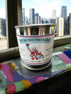 A candle with a label featuring the Statue of Liberty and the text 'Hate Will Never Make Us Great.' The candle is placed on a windowsill with a cityscape in the background.