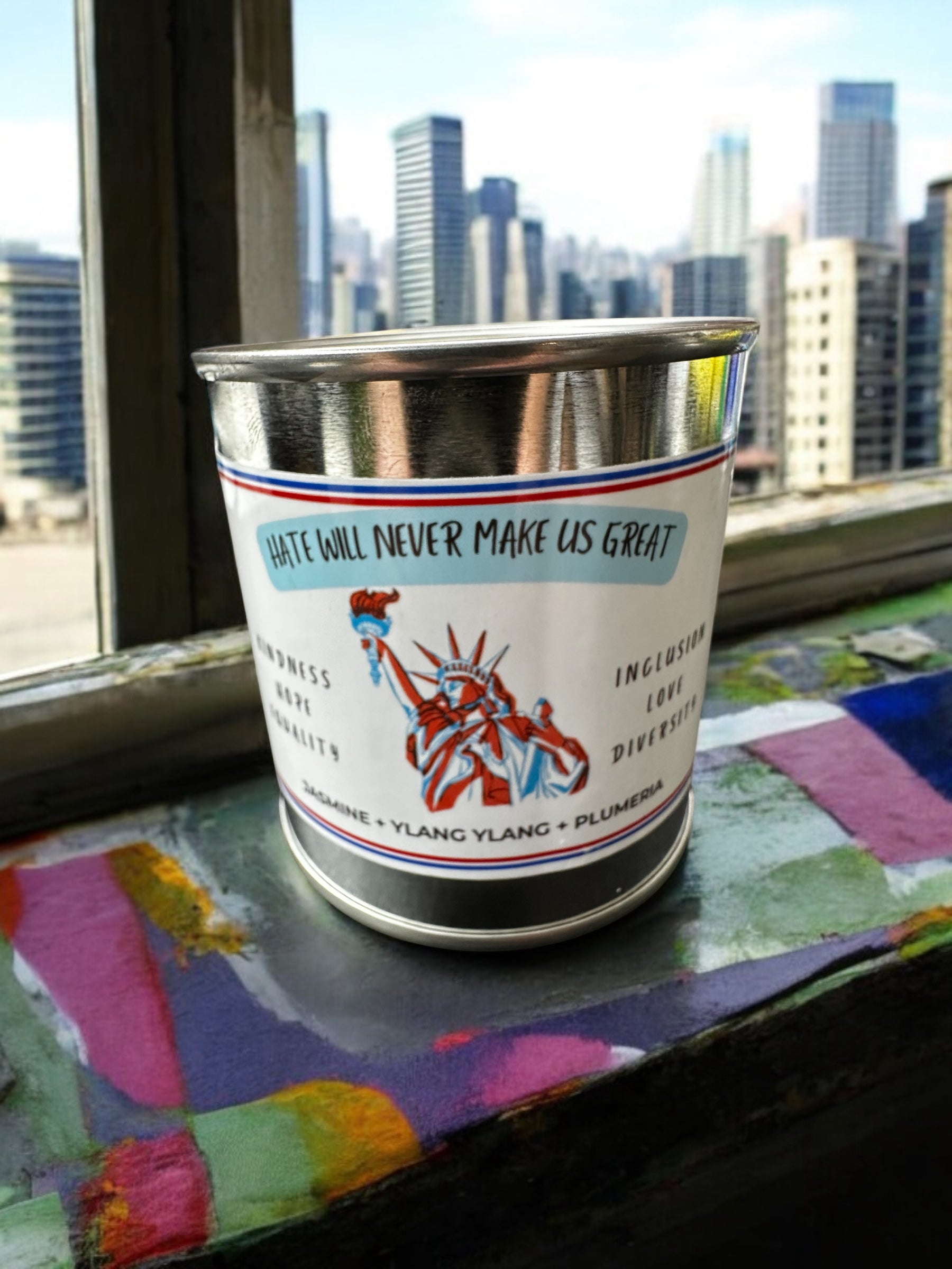 A candle with a label featuring the Statue of Liberty and the text 'Hate Will Never Make Us Great.' The candle is placed on a windowsill with a cityscape in the background.