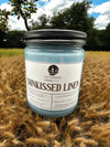 A blue candle jar with a label reading 'Sunkissed Linen', placed in a field with a clear sky in the background.