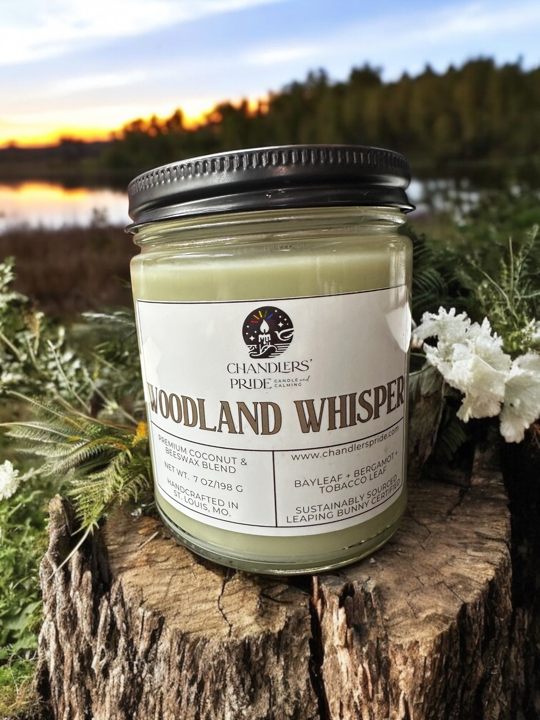 A jar candle with the label 'Woodland Whisper' set on a wooden surface with a backdrop of a serene outdoor scene at sunset.