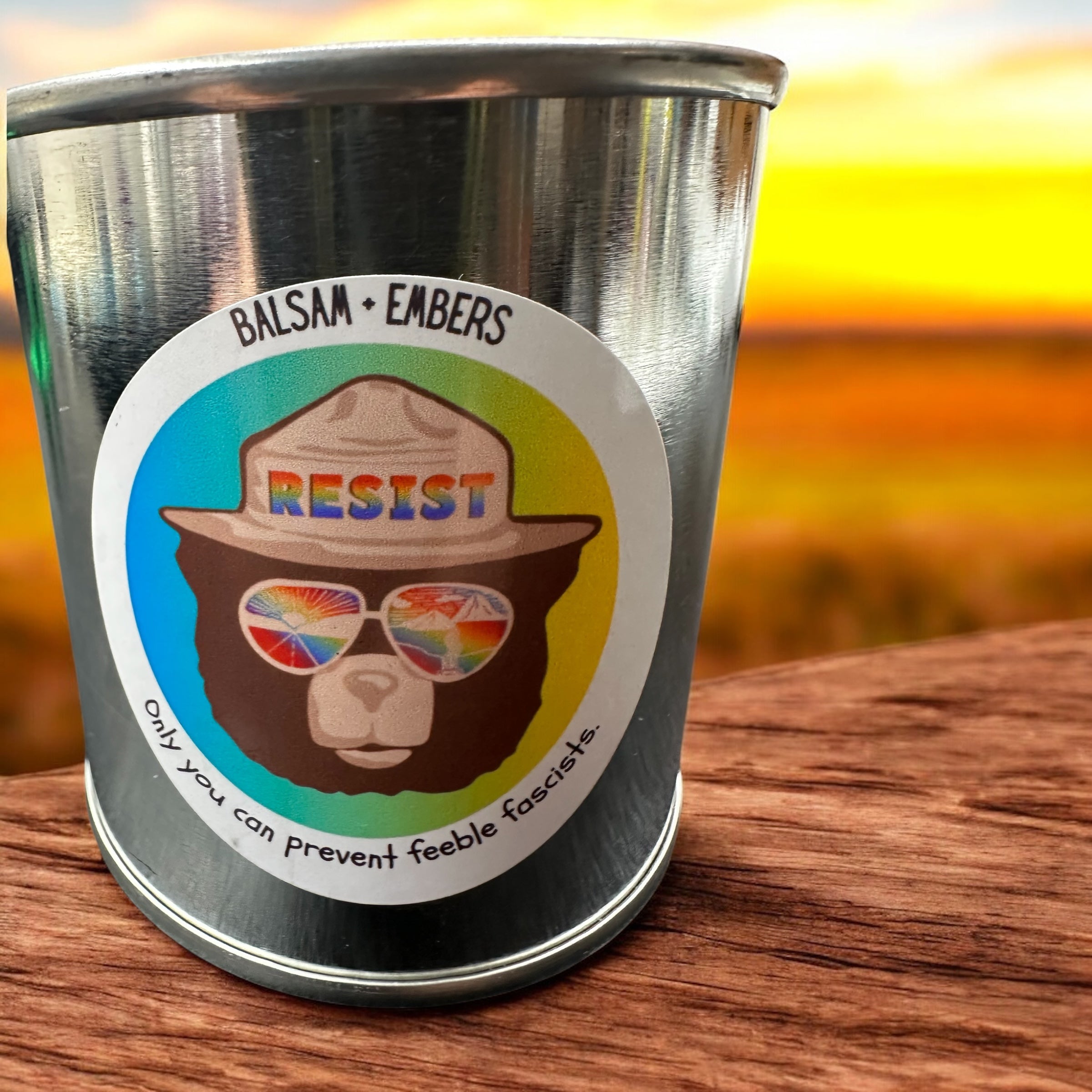 Metal paint can candle with a colorful sticker featuring a bear wearing sunglasses and text on a wooden surface.