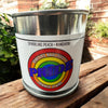 Metal paint can candle with rainbow-colored text and branding on a wooden surface