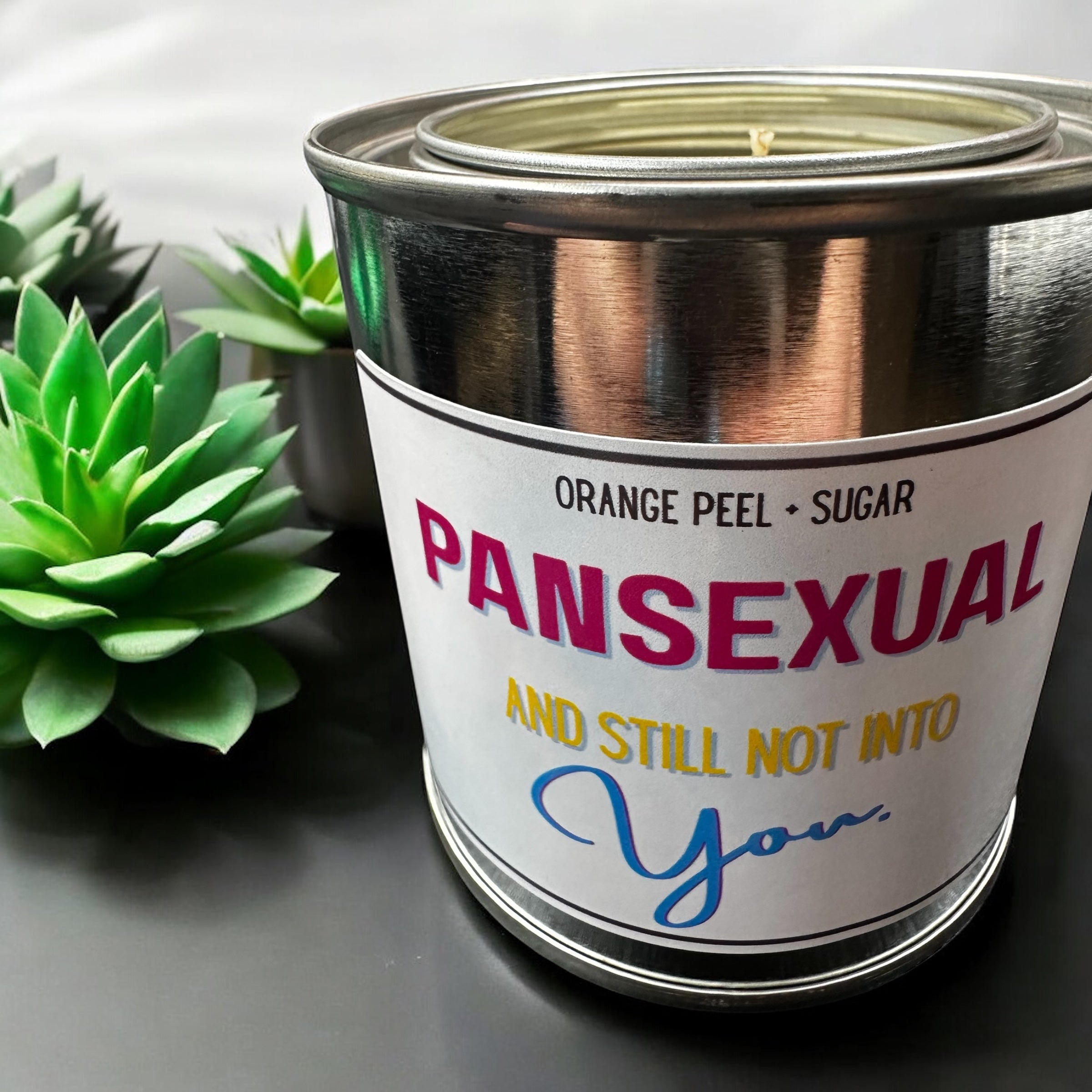 Candle in a metal can with a humorous label next to succulents