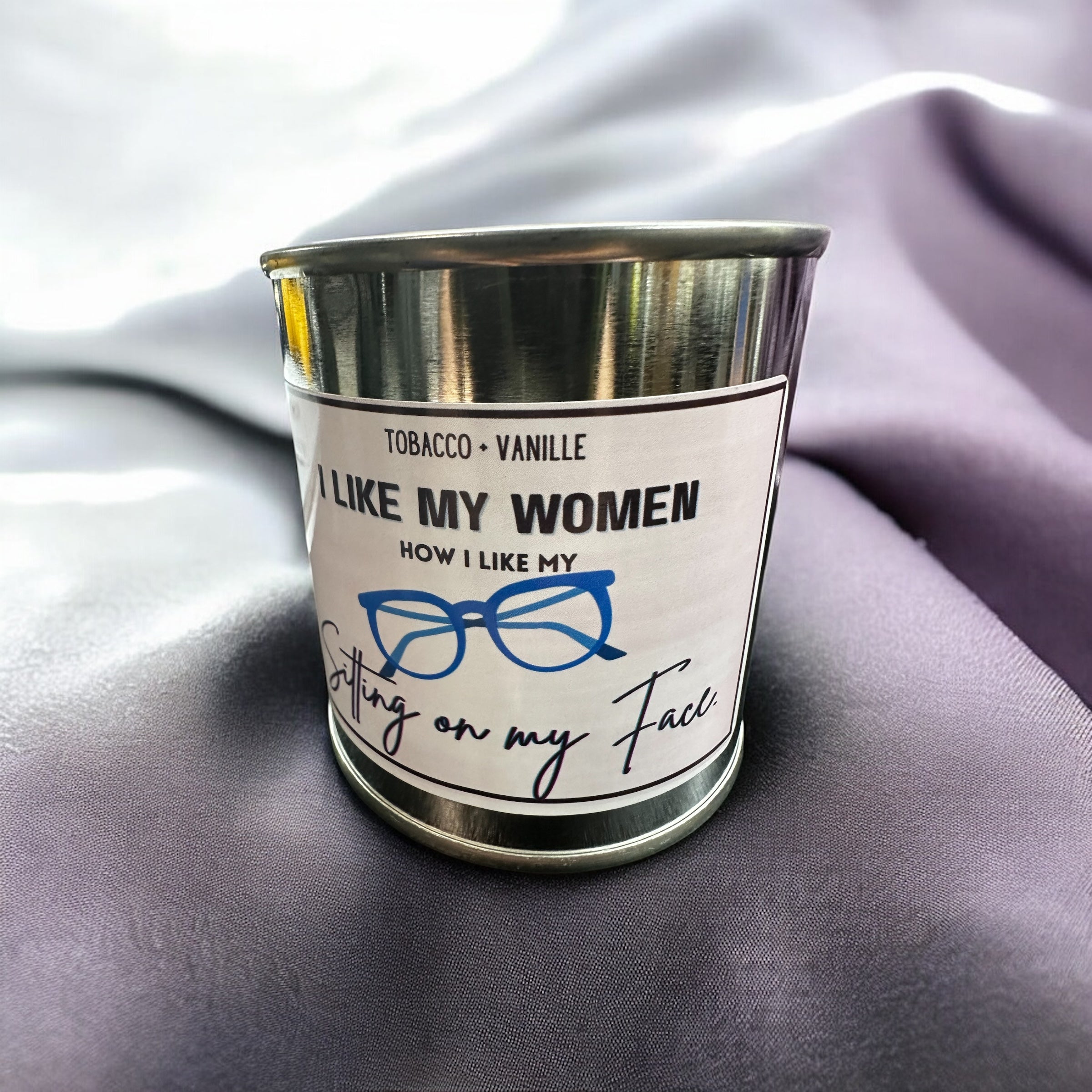 Candle in a tin with a humorous label on a purple fabric background