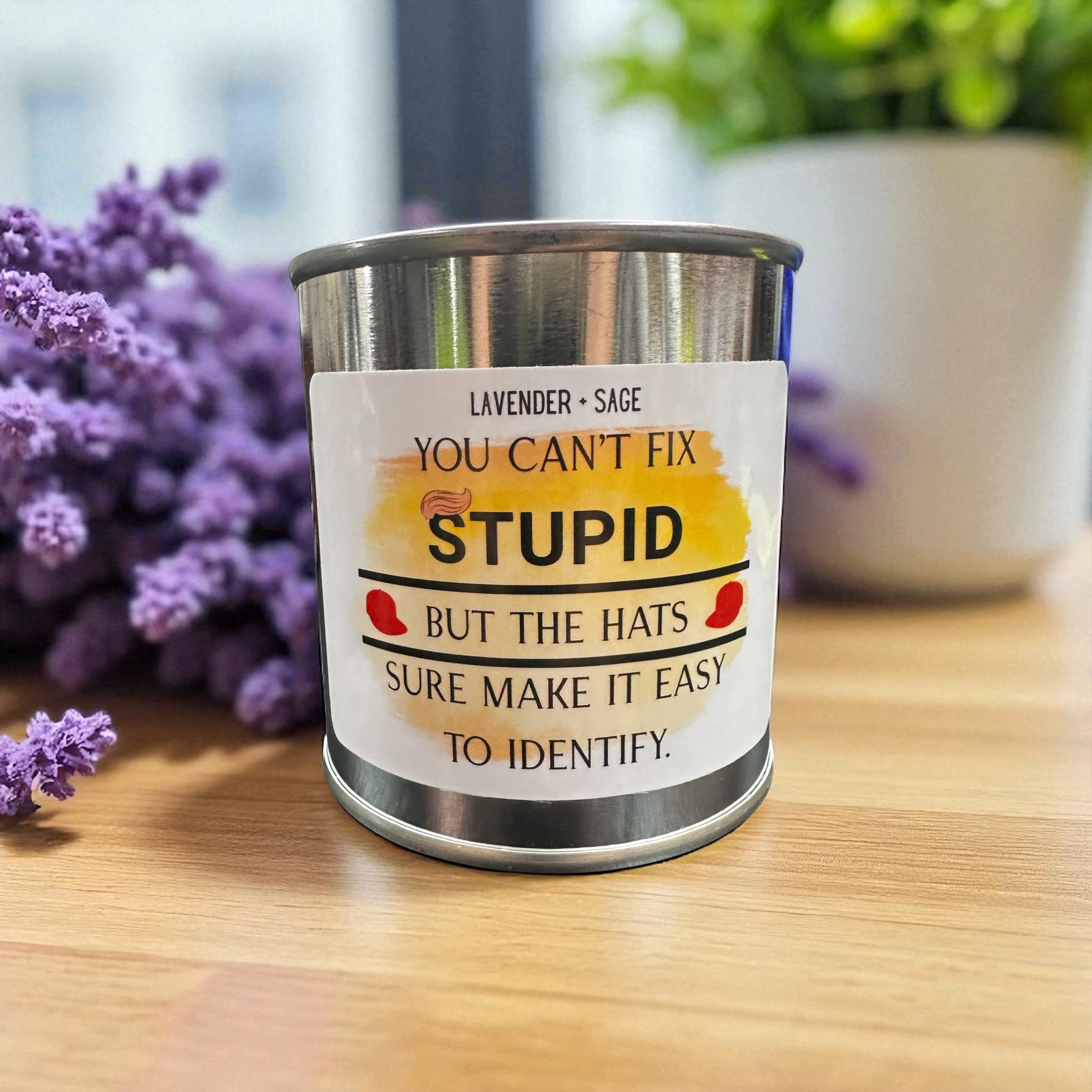 Metal paint can candle with a humorous label on a wooden surface with flowers in the background