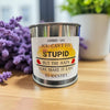Metal paint can candle with a humorous label on a wooden surface with flowers in the background
