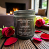 Candle labeled 'Adore' with red roses and petals on a wooden surface.