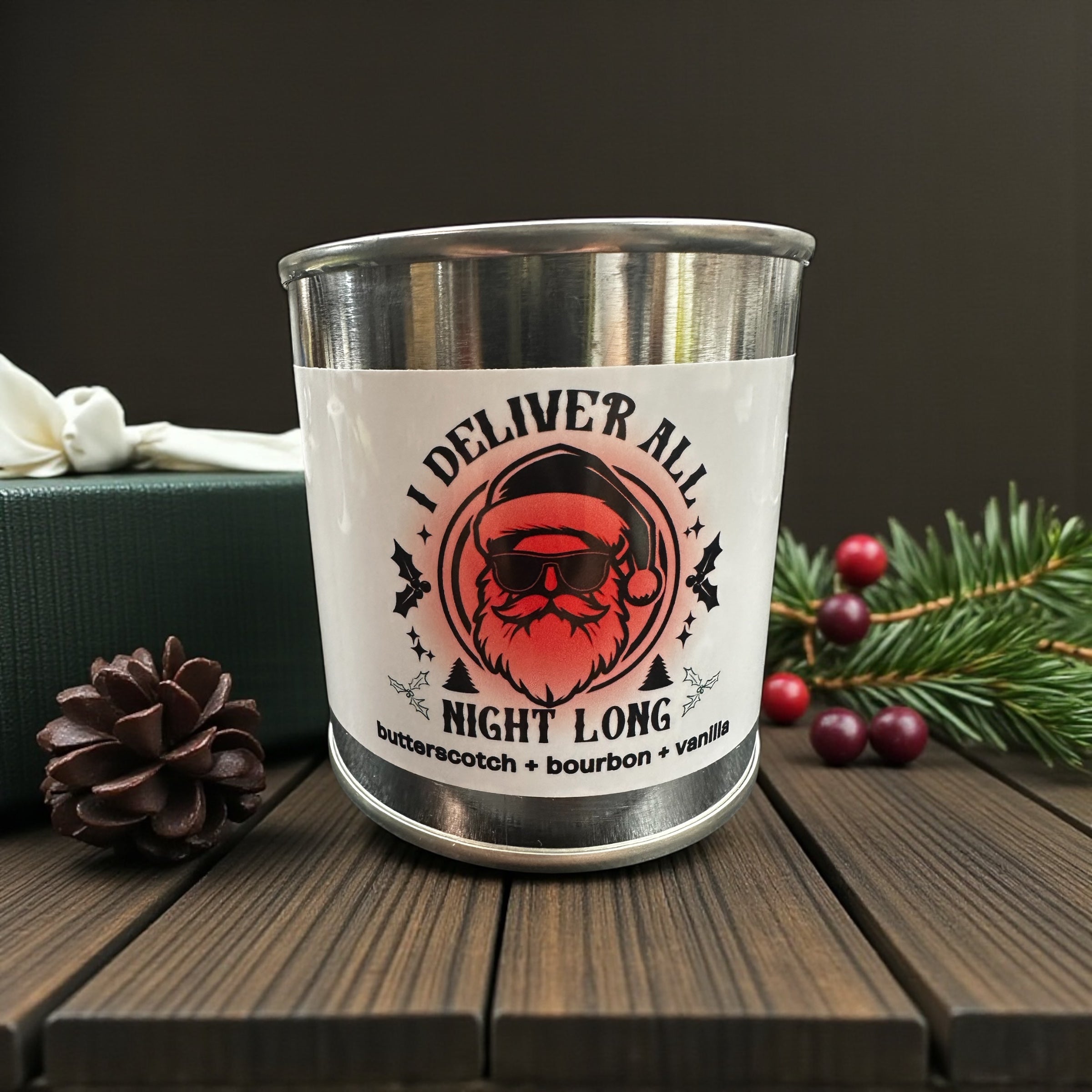 Stainless steel paint can candle with a Santa Claus design and text on a wooden surface with Christmas decorations.